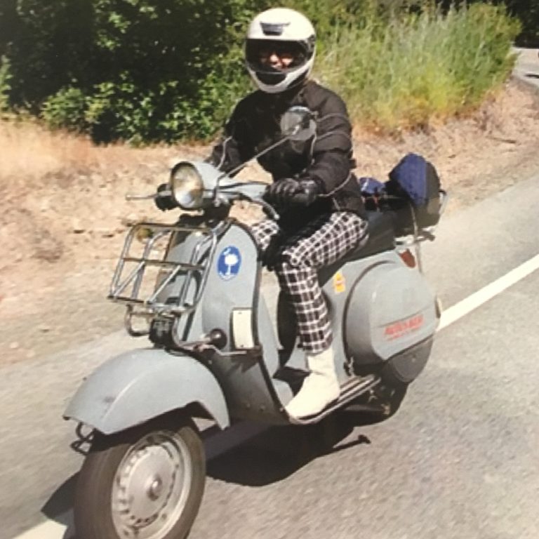 Wearing a full face helmet, riding gear, and plaid pants, Scooterella founder Kate Dana rides a 1978 Vespa P200 around Lake Tahoe, photo ©Kate Dana 2009 