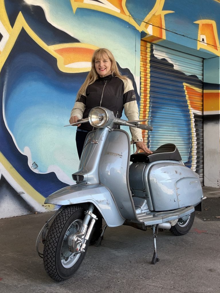 Scooterella founder Kate Dana stands in riding gear beside a light blue, 1969 Lambretta Li125 next to a colorful mural, photo ©Kate Dana