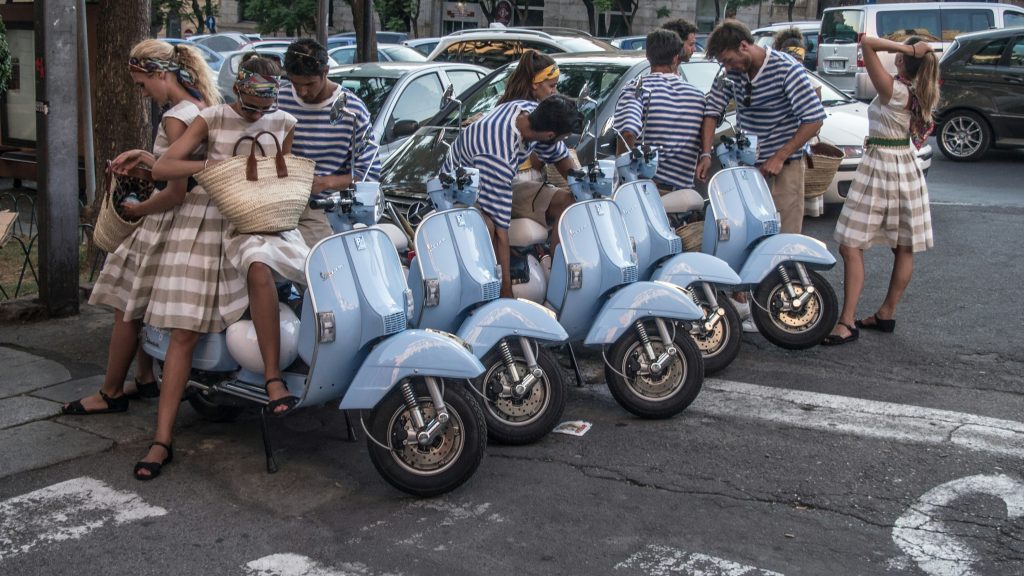 Models in tan clothing gather around several blue vintage Vespa Italian motorscooters in a photo by Ante Hamersmit for unsplash.com