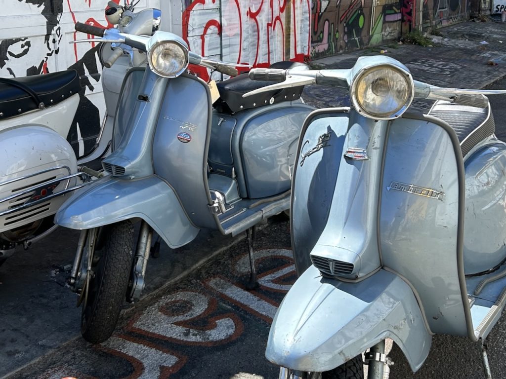 Two silver-blue vintage Lambretta scooters parked on a city street near a graffiti mural, photo ©Kate Dana