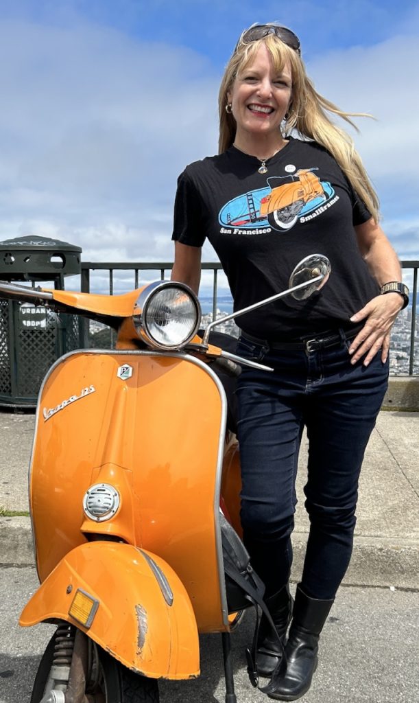 Scooterella founder Kate Dana stands next to an orange 1973 Vespa Primavera in San Francisco, photo ©Kate Dana 2024