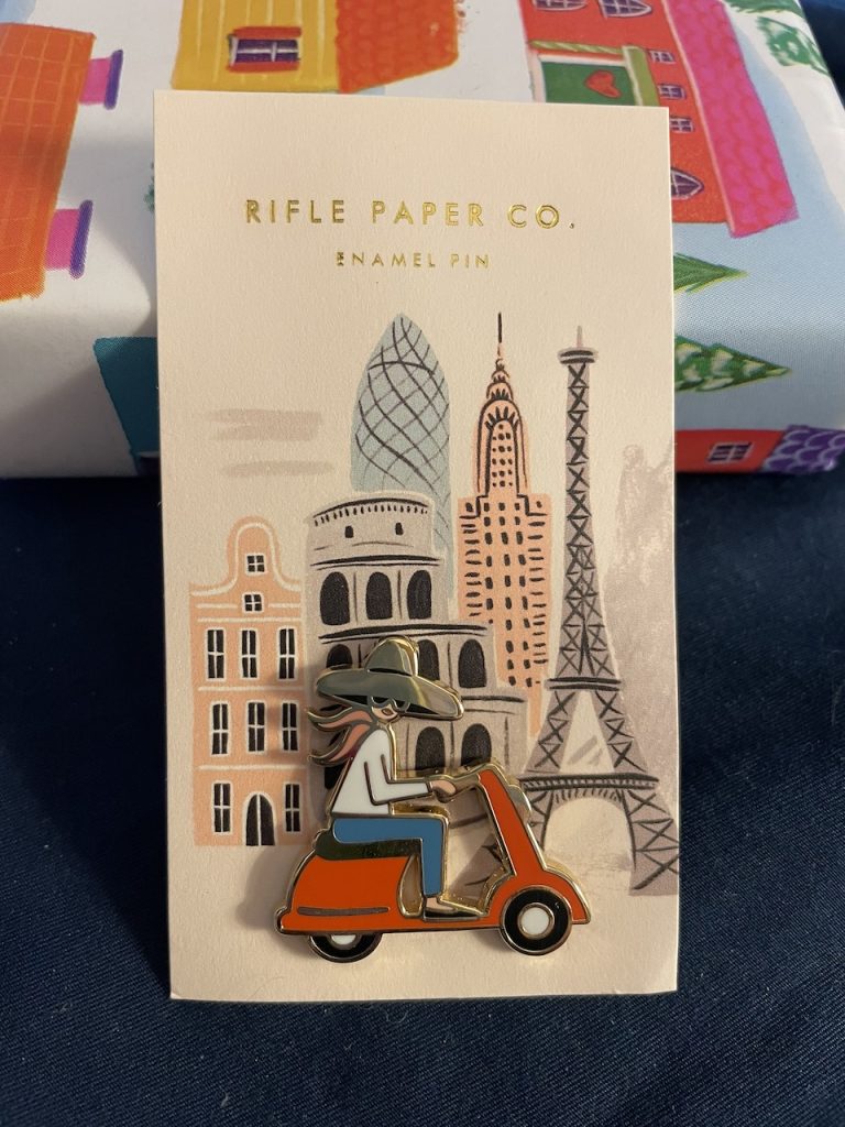 A small lapel pin of a woman riding a scooter on a card with the Paris skyline, photo ©Kate Dana