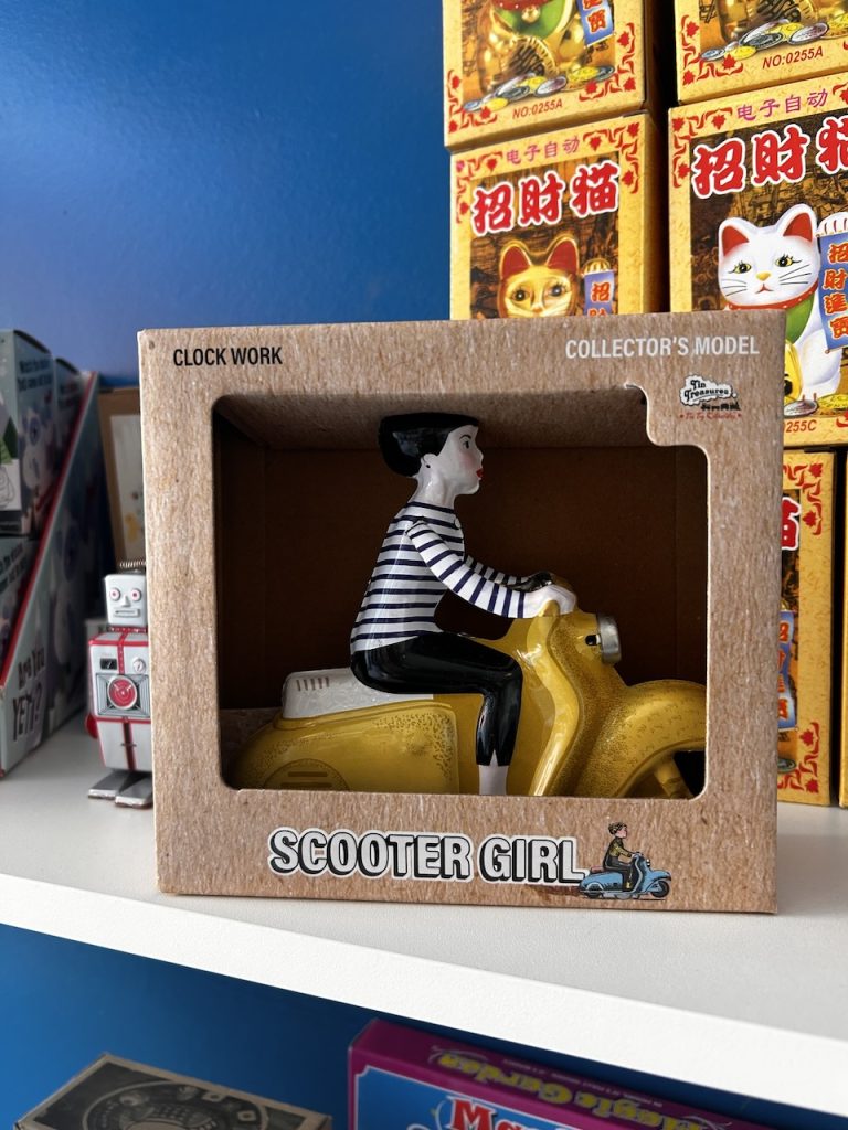 A tin toy featuring a girl with black hair and a striped shirt riding a yellow scooter, photo ©Kate Dana